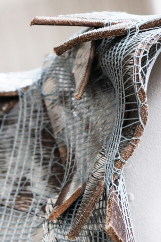 Nahaufnahme von geschichteten, gezackten Stücken aus braunem Holz, die vor einem neutralen Hintergrund mit einem locker gewebten, weißen Netz- oder Maschenmaterial verflochten sind.