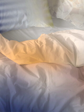 Ungemachtes Bett mit weißen Laken und Kissen, sanft beleuchtet durch natürliches Licht in warmen und kühlen Tönen.