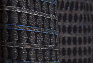 Nahaufnahme eines schwarzen Stoffes mit einem strukturierten geometrischen Muster aus erhabenen, kreisförmigen Formen und dünnen, horizontal verlaufenden blauen Linien.