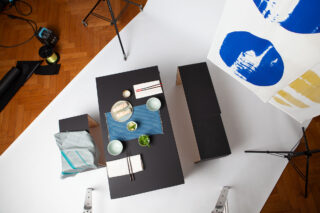 Draufsicht auf einen schwarzen Tisch mit Geschirr, Essen und Essstäbchen vor einem weißen Hintergrund in einem Fotostudio mit Hockern, Lichtern und einem blau-gelben Kunstwerk in der Nähe.