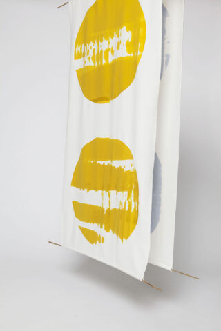 Zwei weiße Stoffbahnen hängen vertikal und jede davon zeigt große Kreisformen mit gelben und grauen abstrakten Mustern. Dünne Holzstangen stützen die Ober- und Unterkante.