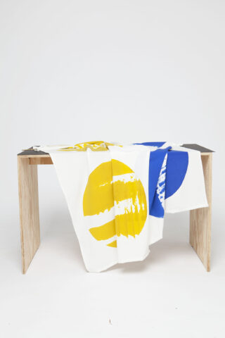 Eine weiße Tischdecke mit großen gelben und blauen kreisförmigen Aufdrucken liegt über einem einfachen Holztisch vor einem schlichten weißen Hintergrund.