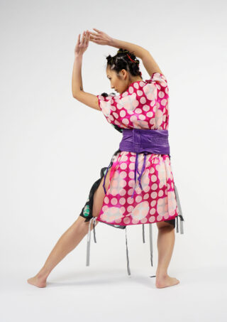 Eine Person, die ein rosa gepunktetes Kleid mit einer violetten Schärpe trägt, posiert mit erhobenen Armen und gebeugten Knien und blickt von der Kamera weg vor einem schlichten weißen Hintergrund.