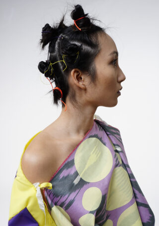Eine Frau mit vier kleinen, gedrehten Dutts, die mit bunten Bändern zusammengehalten werden, und einem schulterfreien Kleidungsstück mit einem kräftigen, kreisförmigen Muster posiert im Profil vor einem schlichten Hintergrund.