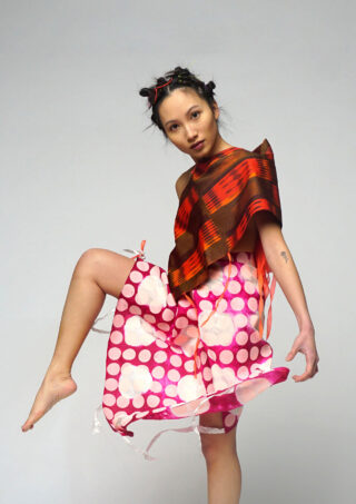 Eine Person posiert mit einem angehobenen Bein und trägt ein rot kariertes Oberteil und einen rosa gepunkteten Rock mit weißen Blumen vor einem schlichten Hintergrund.