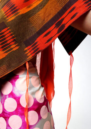 Eine Person trägt ein rot-braun gemustertes Oberteil mit losen orangefarbenen Bändern und einen Rock mit großen rosa-weißen Punkten auf violettem Hintergrund.