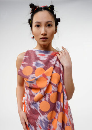 Vor einem schlichten Hintergrund steht eine Frau mit frisiertem Haar. Sie trägt ein gemustertes Kleid mit großen orangefarbenen Punkten und abstrakten Mustern in Rot, Lila und Weiß.