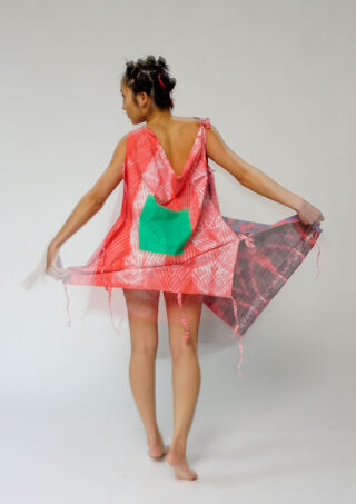 Eine barfuß stehende Person mit abgewandtem Gesicht trägt ein rot-weiß gemustertes Kleid mit grünem Flicken und hält einen Teil des Kleidungsstücks hoch. Der Hintergrund ist schlicht und hell.