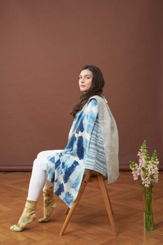 Eine Frau in weißen Leggings, einem blau-weißen Batikschal und gelben Socken sitzt seitlich auf einem Holzstuhl vor einem braunen Hintergrund, auf dem Boden steht eine Vase mit Blumen.