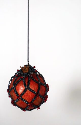 Ein roter Granatapfel, umhüllt von einem dekorativen Netz aus schwarzen und roten Perlen, hängt an einer schwarzen Schnur vor einem schlichten weißen Hintergrund.