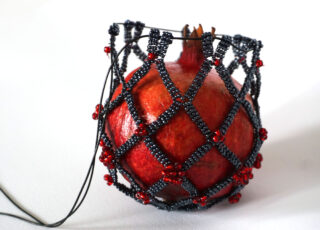 Ein ganzer Granatapfel ist in ein dekoratives Netz aus schwarzen und roten Perlen gehüllt und liegt auf einer weißen Oberfläche.