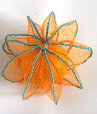 Ein orangefarbenes Objekt, das in ein orangefarbenes Netz eingewickelt und mit blauem Faden umrandet ist, sodass die Form eines Sterns oder einer Blume entsteht und auf einer weißen Oberfläche platziert ist.