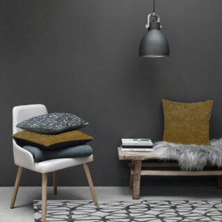 Ein modernes Interieur mit einem weißen Stuhl voller Kissen, einer Bank mit Büchern, einem Kissen und einer Pelzdecke, alles vor einer dunkelgrauen Wand und einer Hängelampe darüber.