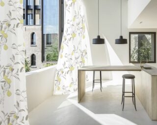 Moderne Küche mit heller Holzinsel, zwei schwarzen Hängelampen und großen Fenstern mit weißen und gelben Blumenvorhängen mit Blick auf Backsteingebäude und Grünflächen.