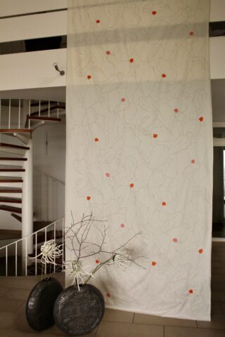 Vor einer Treppe hängt ein hohes weißes Stoffpaneel mit schwachen Strichzeichnungen und verstreuten orangefarbenen und rosa Punkten; darunter stehen zwei Vasen mit kahlen Zweigen.