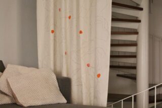 Ein graues Sofa mit zwei beigefarbenen Kissen steht vor einem weißen Vorhang mit hellen Mustern und kleinen orangefarbenen Punkten; im Hintergrund ist eine Wendeltreppe zu sehen.
