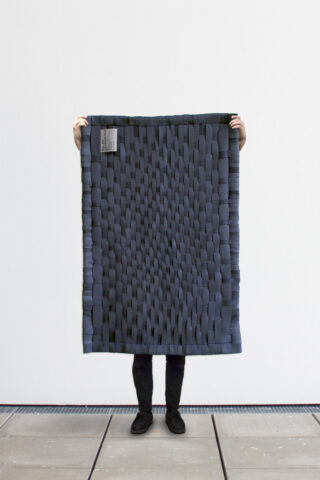 Eine Person steht auf einem gefliesten Boden und hält eine rechteckige, gewebte dunkelgraue Matte mit einem kleinen Etikett in der oberen linken Ecke vor einer schlichten weißen Wand hoch.