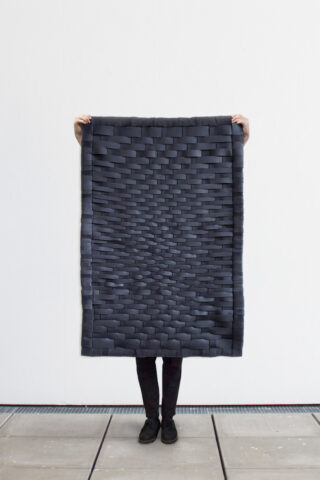 Eine Person steht vor einer weißen Wand und hält eine rechteckige, schwarz gewebte Matte hoch, die ihren Körper bedeckt. Nur ihre Hände und Unterschenkel sind sichtbar.