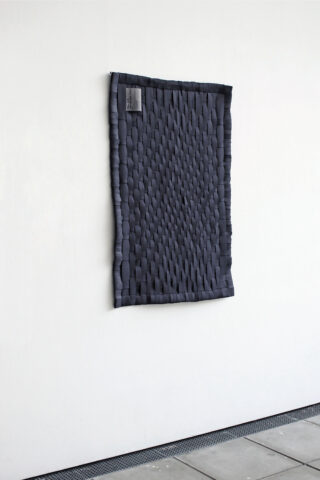 An einer weißen Wand hängt ein rechteckiger, dunkelgrauer gewebter Stoff mit einem kleinen grauen Etikett in der oberen linken Ecke.