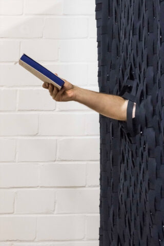 Der Arm einer Person ragt durch eine Lücke in einem dunklen gewebten Paravent und hält ein geschlossenes Buch mit blauem Einband vor einer weißen Ziegelwand.