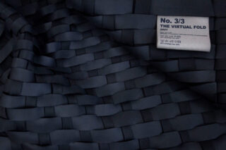 Dunkel gewebter Stoff mit einem weißen Etikett mit dem Text „Nr. 3/3 THE VIRTUAL FOLD 2021“ und Pflegesymbolen, das in der oberen rechten Ecke am Stoff angebracht ist.