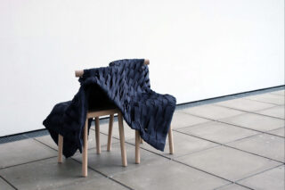 Ein Holzhocker mit hellen Beinen ist mit einem dunklen, gewebten Stoff bedeckt und steht auf einem gefliesten Boden vor einer schlichten weißen Wand.