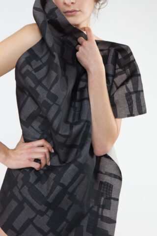 Eine Person hält ein Stück halbtransparenten, schwarz gemusterten Stoff hoch, das über ihre Schulter drapiert ist und ihren Oberkörper teilweise bedeckt.