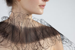 Eine Person trägt einen transparenten, schwarzen Kragen mit komplizierten, geschwungenen Fadenmustern, der von den Schultern aufwärts vor einem schlichten Hintergrund zu sehen ist.