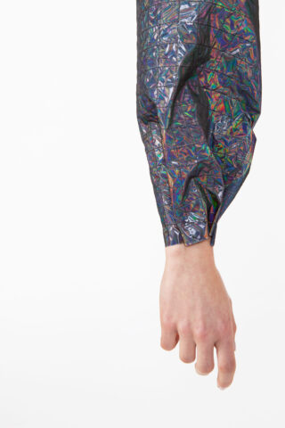 Der Arm einer Person in einem glänzenden, zerknitterten, mehrfarbigen Ärmel, die Hand entspannt vor einem weißen Hintergrund.