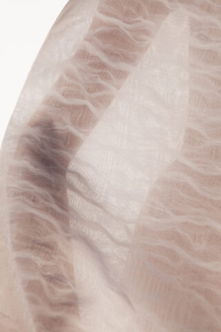 Ein transparenter, strukturierter Stoff mit einem Wellenmuster ist über den Arm einer Person drapiert, wirft weiche Schatten und enthüllt verschwommene Hauttöne darunter.