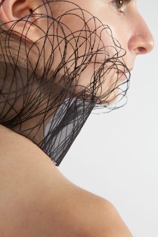 Nahaufnahme des Gesichts und der Schulter einer Person mit einem dekorativen Netzkragen mit geschwungenen schwarzen Linien um den Hals.