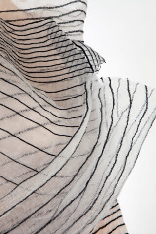 Nahaufnahme eines durchsichtigen, weißen, leichten Stoffes mit schwarzen horizontalen und diagonalen Streifen, der vor einem schlichten Hintergrund ein vielschichtiges und fließendes Erscheinungsbild erzeugt.