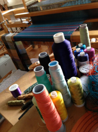 Spulen mit buntem Garn und Fäden sind auf einem Tisch vor einem Webstuhl mit einem teilweise gewebten Textil angeordnet.