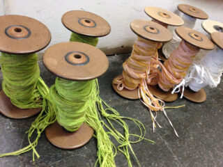 Mehrere Holzspulen, die mit grünen, gelben, pfirsichfarbenen und weißen Fäden umwickelt sind, sind auf einer dunklen Oberfläche vor einer hellen Wand angeordnet.