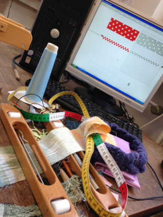 Ein hölzerner Webschützen, Maßbänder und Nähmaterialien auf einem Schreibtisch vor einem Computer, auf dem Webmuster angezeigt werden.