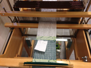 Nahaufnahme eines Webstuhls mit weißen Kettfäden und teilweise eingewebtem grün-schwarz gemustertem Stoff. Auf dem Stoff liegt ein Blatt Papier.