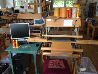 Ein Webstuhl mit orangefarbenen und gelben Fäden, ein Computerarbeitsplatz und in einem gut beleuchteten Studio ein gewebter Stoff in Arbeit.