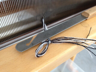 Eine Nahaufnahme eines Webstuhls mit einem Einfädelwerkzeug aus Metall und schwarz-weißem Garn, das durch das Webblatt des Webstuhls gefädelt ist.