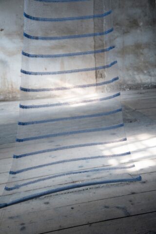 Ein durchsichtiger Stoff mit horizontalen blauen Streifen hängt vor einer strukturierten, verwitterten Wand und einem Holzboden, beleuchtet von natürlichem Licht.