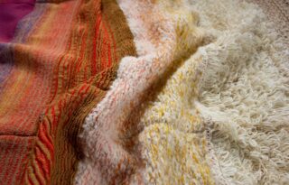 Eine Nahaufnahme einer strukturierten, mehrfarbig gewebten Decke mit Abschnitten aus orangefarbenem, rotem, braunem und weißem Garn mit sowohl glatten als auch flauschigen Bereichen.