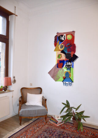 Eine gemütliche Zimmerecke mit einem Holzsessel, einer Topfpflanze, Büchern auf dem Boden, einem bunten gewebten Wandbehang und einem großen Fenster, das natürliches Licht hereinlässt.