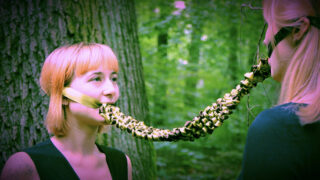 Zwei Menschen stehen sich in einem Wald gegenüber, verbunden durch ein dekoratives, gold-schwarzes Mundstück, das ihre beiden Münder bedeckt.