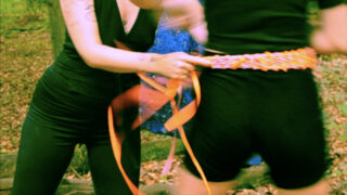 Zwei Personen draußen, eine löst oder zieht ein orangefarbenes Band um die Taille der anderen, im Hintergrund sind abgefallene Blätter und Bäume zu sehen.