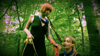 Zwei Frauen in schwarzen Outfits stehen in einem Wald, jede mit farbigen Seilen im Mund und um den Hals, und halten Händchen.
