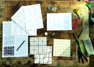 Ein Holzschreibtisch mit ordentlich angeordneten Zeichnungen geometrischer Muster, Bleistiften, Scheren, einem Lineal, einem Radiergummi, einem Spitzer, einem Textmarker und einem Federmäppchen.