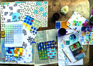 Eine Tischplatte mit Aquarellgemälden geometrischer Formen, einer Farbpalette, Pinseln, Tassen, einer Kerze und Blättern Papier, angeordnet in einem kreativen Arbeitsbereich.