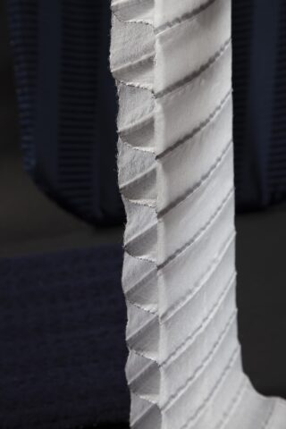 Eine Nahaufnahme eines vertikalen, weißen, plissierten Stoffstreifens mit sichtbaren Nähten vor einem dunklen, unscharfen Hintergrund.