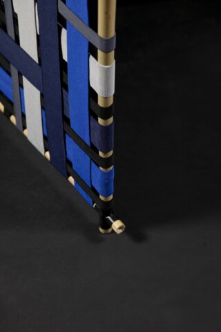 Eine Nahaufnahme einer gewebten Struktur aus blauen, schwarzen und grauen Stoffbändern, die um vertikale und horizontale Goldstäbe auf einem dunklen Hintergrund gewickelt sind.