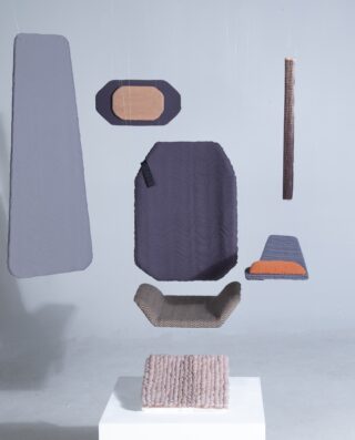 Verschiedene geometrische Stoffpaneele und Kissen in Grau- und Brauntönen hängen an dünnen Drähten über einem weißen Sockel in einem minimalistischen Raum.