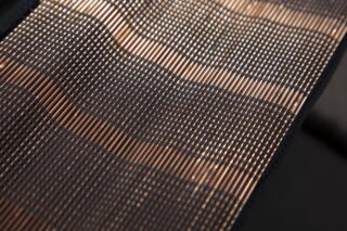 Nahaufnahme einer gewebten Matte mit abwechselnd dunklen und hellen Streifen, die feine, sich kreuzende Fäden und eine strukturierte Oberfläche zeigt.